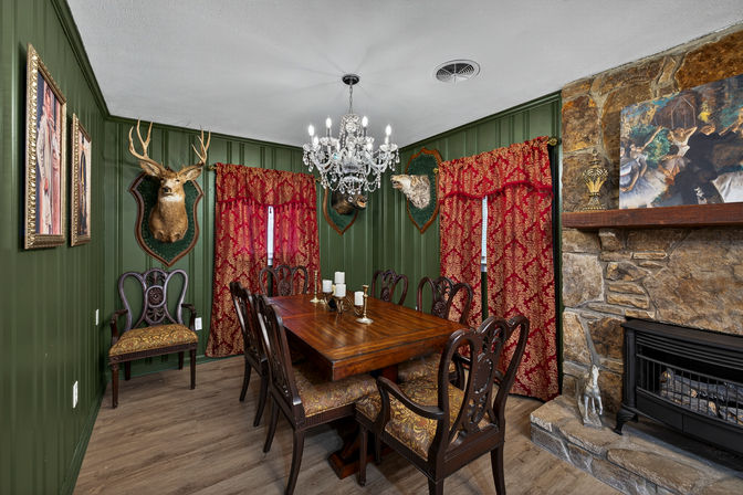 Cozy lodge-style dining room with green paneled walls, mahogany dining table and chairs under a crystal chandelier, red damask curtains, mounted deer and animal heads, and a stone fireplace.