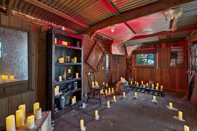Rustic wood-paneled cabin interior with corrugated metal ceiling, scattered flameless candles, an antique chair, bookshelf and stacked books under moody red lighting.