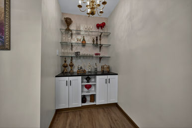 Cozy home bar nook in an alcove with black countertop and white cabinets, three glass shelves holding glassware and gold decorative accents under a brass chandelier