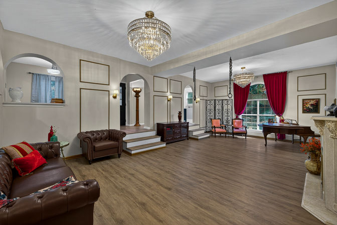 Spacious luxury living room interior with crystal chandeliers, brown leather Chesterfield sofas, wood-look flooring, arched doorways, red velvet curtains framing a large window, grand piano and decorative fireplace.