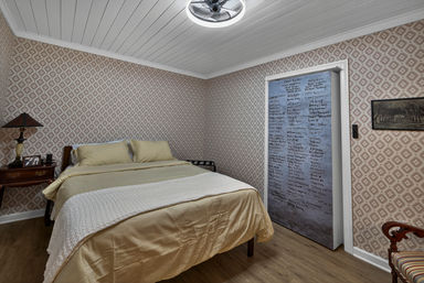 Cozy guest bedroom with beige bedding and white waffle throw, pink geometric wallpaper, wooden nightstand with lamp and framed photo, hardwood floor, ceiling fan light, and a sliding closet door covered in handwritten names.