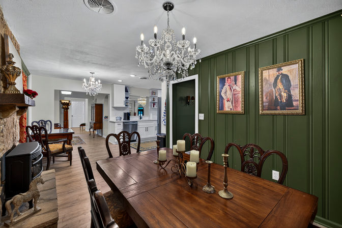 Traditional dining room with large wooden table and candles, green paneled accent wall with framed portraits, crystal chandeliers and open-concept white kitchen in background