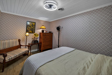Cozy vintage-style bedroom with geometric patterned wallpaper, striped wooden settee, antique gramophone on a side table, tall chest of drawers, standing coat rack, warm lamps, and a cream-covered bed.