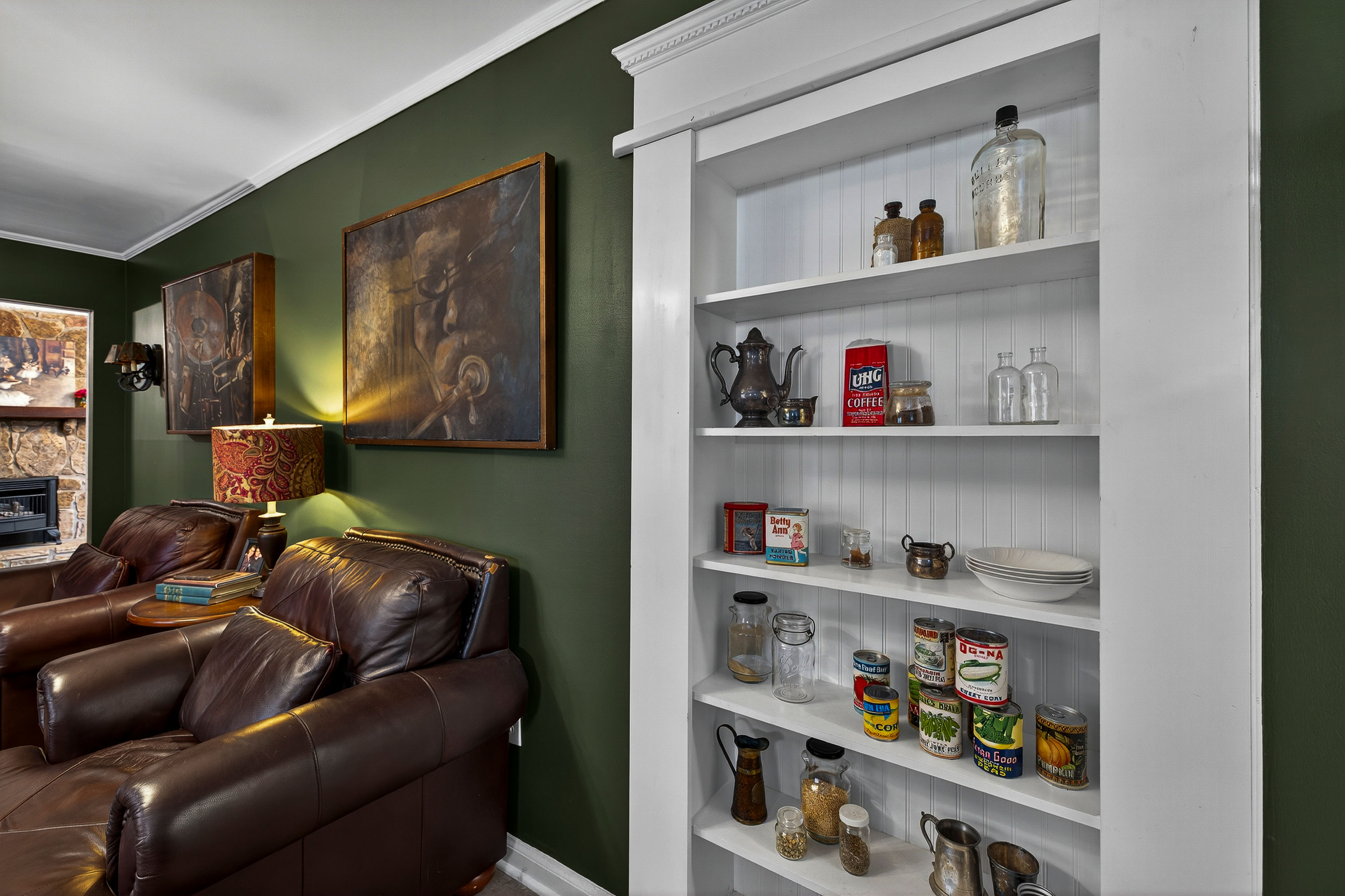 Cozy living room interior with dark green wall, two brown leather armchairs by a patterned lamp and framed artwork, and a white built‑in shelf displaying vintage tins, glass jars, plates and metal teapots, with a stone fireplace visible at left.