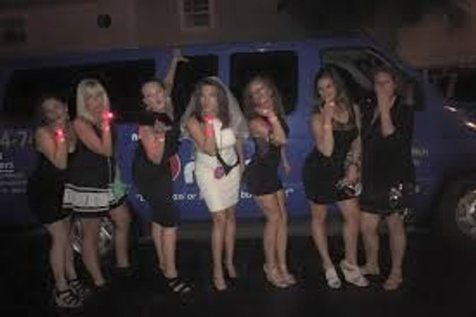 Bachelorette party of seven women posing on a city street at night in front of a blue party van, bride in a white dress and veil surrounded by friends in black dresses wearing glowing red wristbands.
