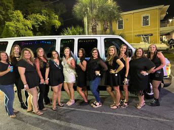 Smiling bachelorette party group of women, one in a white sash and dress, posing with drinks in front of a white party van at night by palm trees and a marina-style building.