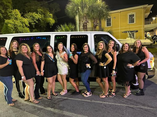 Smiling bachelorette party group of women, one in a white sash and dress, posing with drinks in front of a white party van at night by palm trees and a marina-style building.