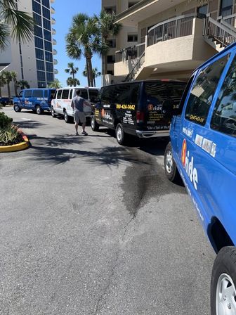 Beachfront condo shuttle vans lined up under palm trees on a sunny day — blue and black passenger vans parked along a driveway with a person walking between them.