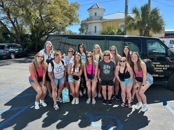 Cheerful group of women and teenage girls posing in front of a black shuttle van in a sunny Florida beach-town parking lot with palm trees and coastal-style buildings.