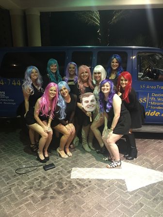 Group of women in colorful wigs and black outfits posing at night in front of a blue party van, one holding a large cutout face — festive bachelorette/party vibe outside a venue.