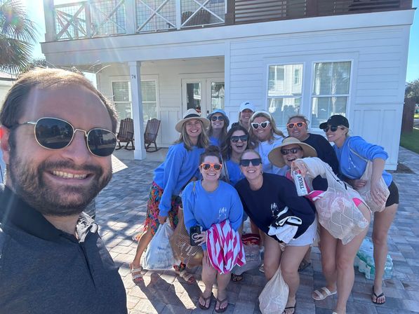 Smiling group selfie outside a white beach house — friends in sunglasses, sun hats and blue shirts holding towels and mesh beach bags on a sunny coastal getaway.