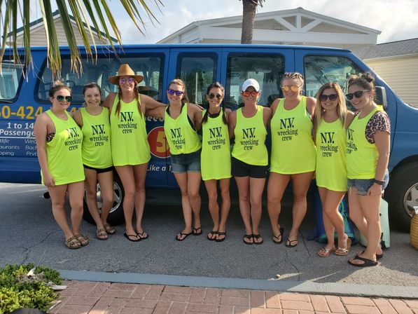 Sunny beachside group of ten women wearing matching neon yellow "N.V. wrecking crew" tank tops, sunglasses and sandals posing in front of a blue passenger van under a palm tree on a coastal street.