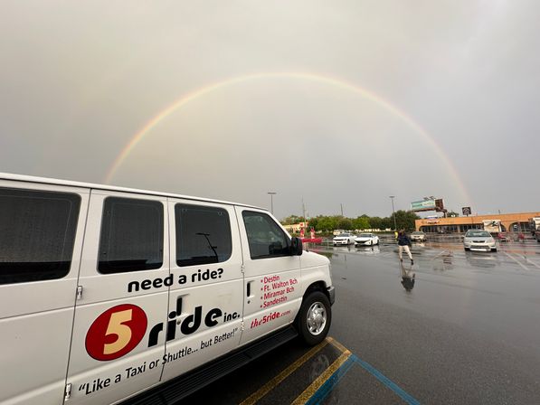 Vibrant rainbow arching over a wet shopping-center parking lot with a white passenger shuttle van in the foreground, puddle-reflections of cars and a person walking.