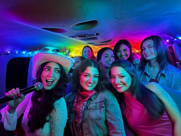 Smiling group of women enjoying karaoke in a neon-lit party van, one holding a microphone and wearing a cowboy hat.