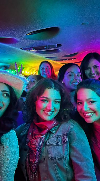 Smiling group of women enjoying karaoke in a neon-lit party van, one holding a microphone and wearing a cowboy hat.