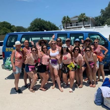 Group of women in matching pink shirts and fanny packs posing and waving on a sunny sandy beach in front of a blue passenger van, with palm trees and a beach house in the background.