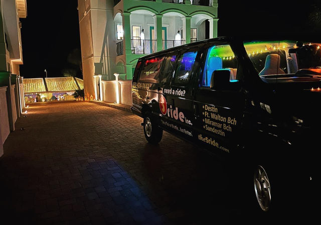 Nighttime coastal resort driveway with a black shuttle van glowing with colorful interior lights parked beside a white balustraded building and string-lit entrance.