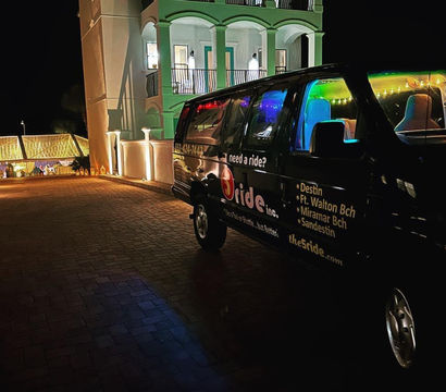 Nighttime coastal resort driveway with a black shuttle van glowing with colorful interior lights parked beside a white balustraded building and string-lit entrance.