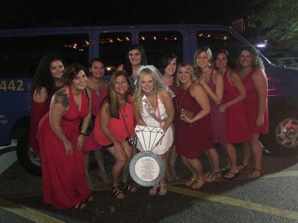 Bride-to-be in a white sash and veil posing with a group of women in red dresses in a nighttime parking lot in front of a blue van — playful bachelorette party photo.