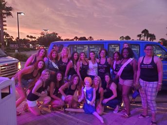 Bachelorette party group of women in matching tank tops and sashes posing with an inflatable doll in front of a blue party van at sunset, palm trees and a pink-purple sky over a coastal parking lot