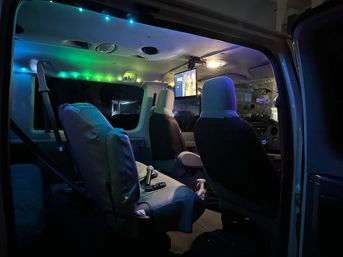 Nighttime camper-van interior lit by blue-green LED strips, two front captain seats, swivel middle seat, ceiling-mounted TV and cozy road-trip setup.