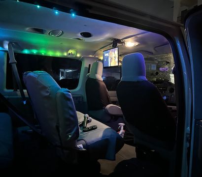 Nighttime camper-van interior lit by blue-green LED strips, two front captain seats, swivel middle seat, ceiling-mounted TV and cozy road-trip setup.