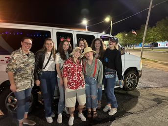 Eight friends in casual and playful outfits (Hawaiian shirt, bucket hats, mouse ears, fake mustache) smiling and posing in front of a white van with purple interior lights in a nighttime parking lot with an American flag in the background.