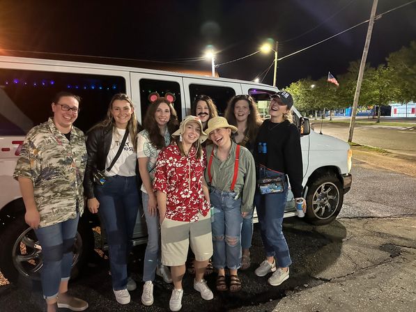 Eight friends in casual and playful outfits (Hawaiian shirt, bucket hats, mouse ears, fake mustache) smiling and posing in front of a white van with purple interior lights in a nighttime parking lot with an American flag in the background.