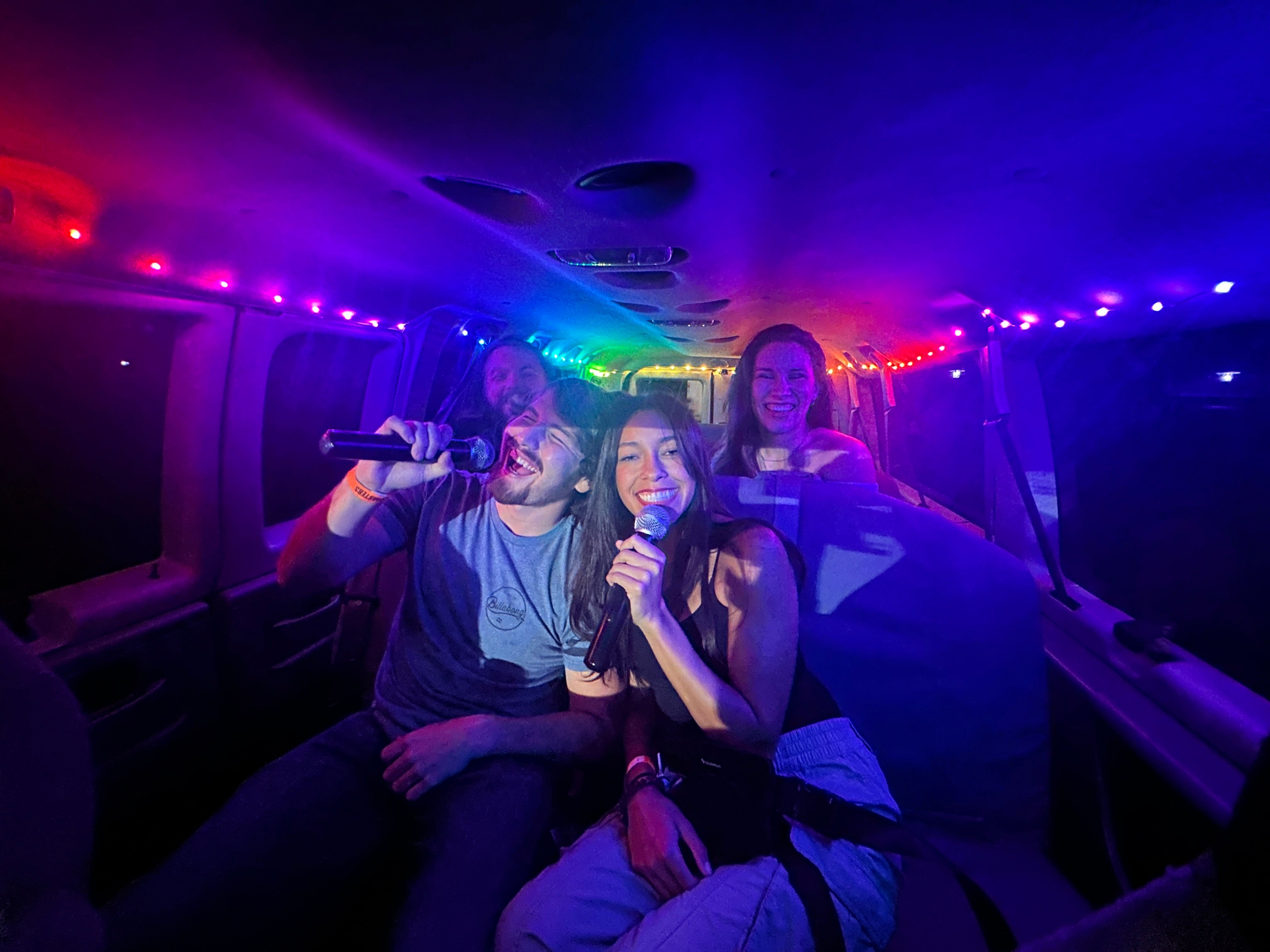 Four friends singing karaoke inside a party van, holding microphones and laughing under rainbow LED string lights during a nighttime ride.