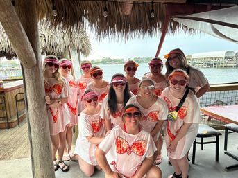 Group of women in matching pink beach cover-ups with orange bikini print and visors posing under a thatched tiki hut by a marina waterfront — playful girls‑trip photo.