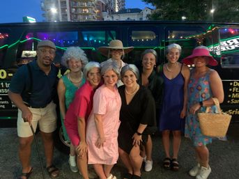 Ten friends in playful elderly-themed costumes — gray wigs, dresses, pearls, hats and a cane — smiling and posing in front of a black party van at dusk on a downtown city street.