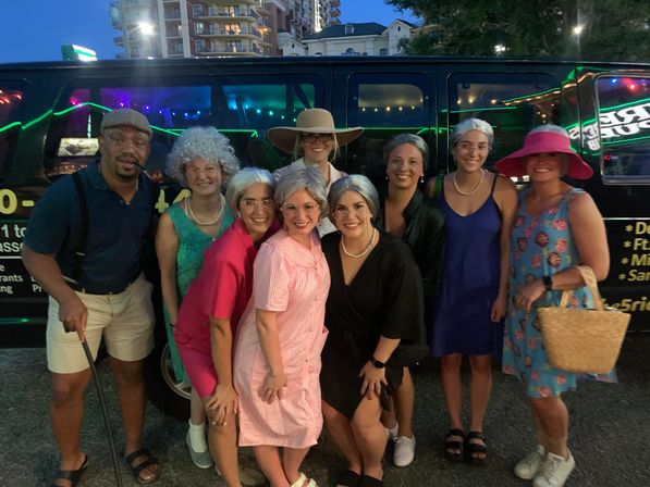 Ten friends in playful elderly-themed costumes — gray wigs, dresses, pearls, hats and a cane — smiling and posing in front of a black party van at dusk on a downtown city street.