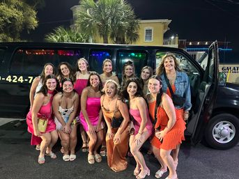 Group of smiling women in colorful party dresses posing in front of a black party van at night with palm trees and lit buildings — friends on a lively coastal night out.