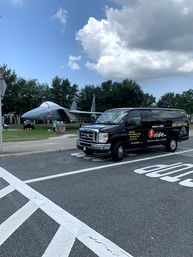 Black passenger shuttle van parked on the road in front of a large gray military jet on display in a grassy outdoor aviation exhibit under a partly cloudy blue sky.
