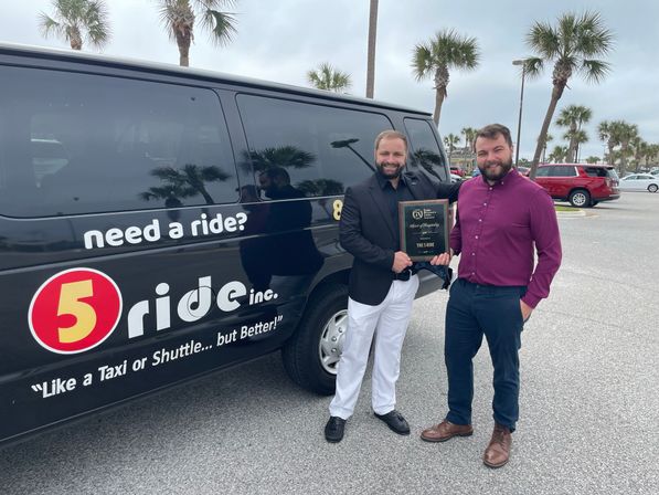 Two men smiling and holding an award plaque beside a black shuttle van with 'need a ride?' text in a palm-tree lined beachside parking lot.