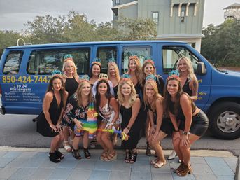 Smiling bachelorette party of about a dozen women in black outfits wearing colorful fiesta tiaras, holding a rainbow piñata and posing in front of a blue passenger shuttle van on a neighborhood street.