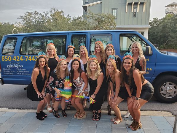 Smiling bachelorette party of about a dozen women in black outfits wearing colorful fiesta tiaras, holding a rainbow piñata and posing in front of a blue passenger shuttle van on a neighborhood street.