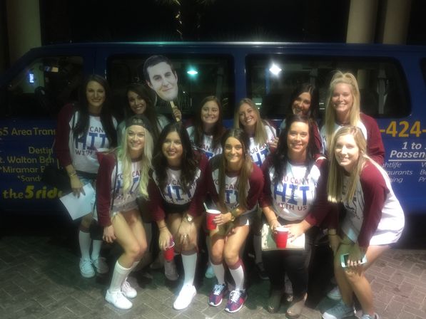 Group of a dozen young women in matching maroon-and-white baseball-style shirts posing at night in front of a blue party van, holding red cups and a large face cutout, smiling for the camera.