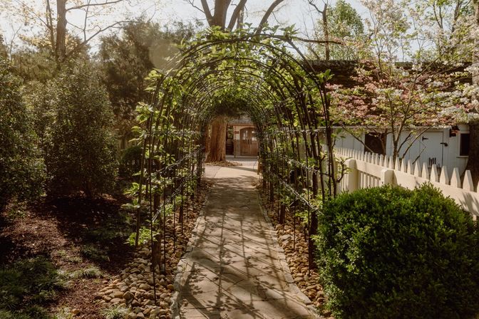Sunlit garden archway with a vine-covered metal trellis over a flagstone path leading to a cottage entrance, framed by a white picket fence, flowering trees, and trimmed shrubs.