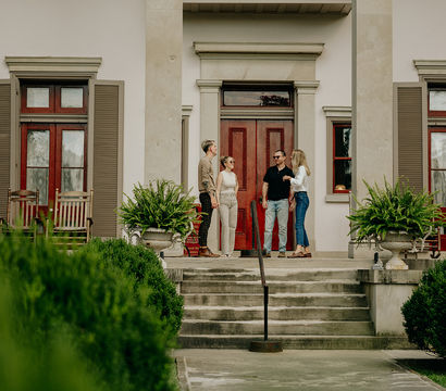 Four people chatting on the stone front steps of a historic house with red double doors, tall columns, wooden shutters, rocking chairs and large fern planters