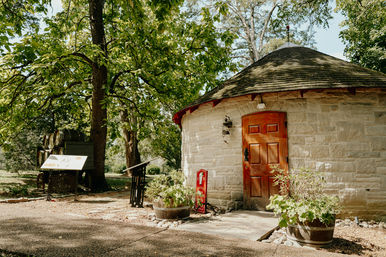 Round historic stone outbuilding with wooden door and shingled roof beside oak trees, barrel planters and interpretive signs along a sunlit garden path