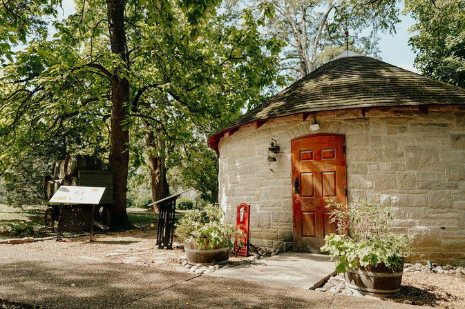 Round historic stone outbuilding with wooden door and shingled roof beside oak trees, barrel planters and interpretive signs along a sunlit garden path