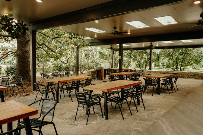 Covered garden patio with wooden tables and black metal chairs, skylights and ceiling fans overhead, stone floor and lush trees—casual outdoor dining terrace.
