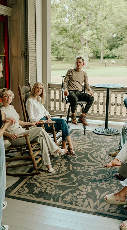 Cozy porch gathering: a woman addresses a group seated in rocking chairs on a covered front porch overlooking a green park-like lawn.