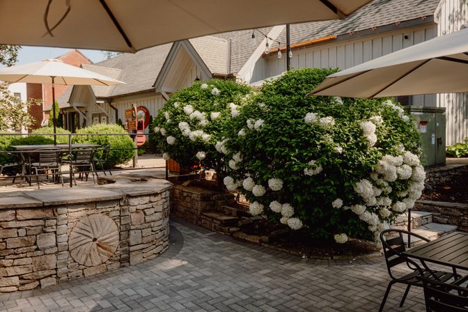 Sunlit outdoor patio with umbrella-shaded metal tables, stone wall featuring a millstone detail, and large white hydrangea bushes by a farmhouse-style building.