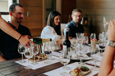 Wine tasting in a winery tasting room — server pouring sparkling wine into a flute as a group enjoys red and white wines and small appetizer plates on a wooden table.