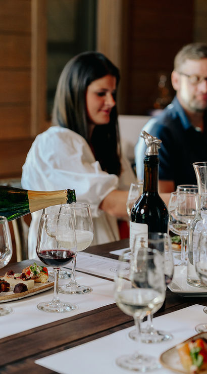 Wine tasting in a winery tasting room — server pouring sparkling wine into a flute as a group enjoys red and white wines and small appetizer plates on a wooden table.
