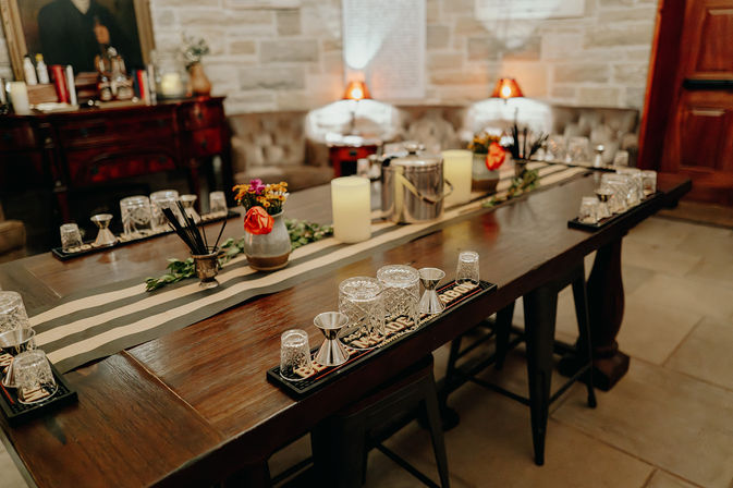 Rustic wooden table arranged for a cocktail tasting with crystal glasses, jiggers, striped runner, candles and small flower vases in a cozy stone‑walled indoor lounge