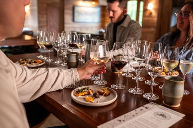 Hand reaching for a glass of white wine amid rows of red and white pours and small appetizer plates on a cozy rustic winery tasting-room bar, people chatting in the background.