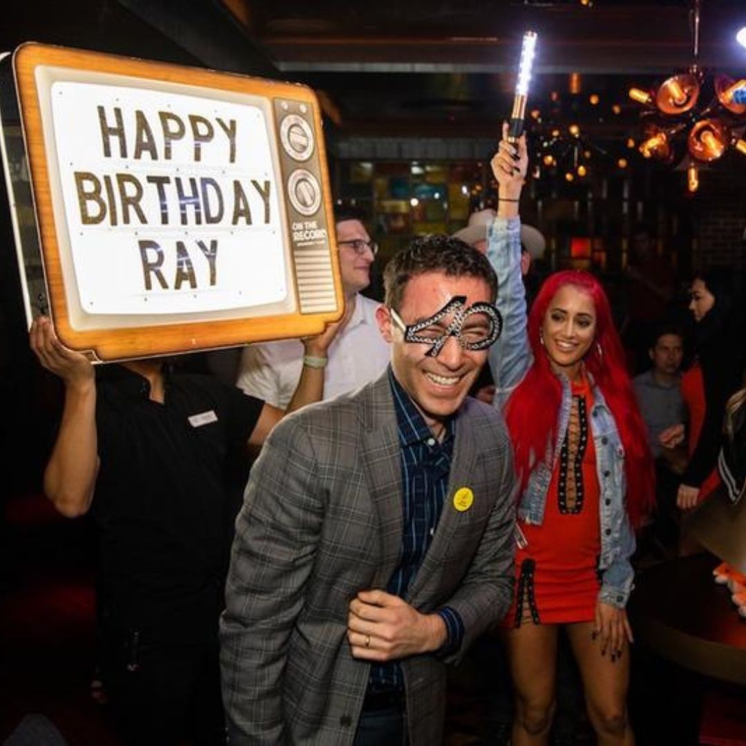 Smiling man in a plaid blazer wearing novelty glitter glasses at an indoor birthday party, LED sparkler and retro TV sign reading 'HAPPY BIRTHDAY RAY' held nearby, woman with bright red hair in a red dress in the background.